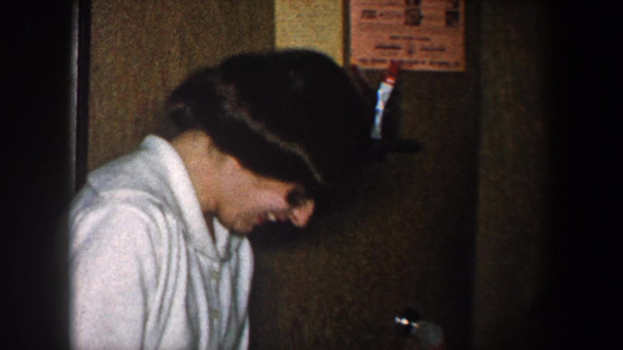 In 1960 New York City home-movie footage, a woman in a white collared shirt laughs in a dim interior near a doorway.