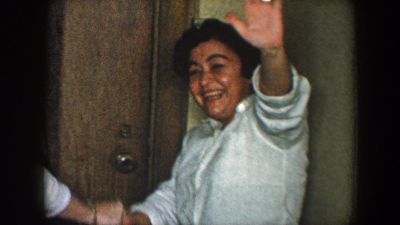 Woman in a white shirt smiles and raises a hand while seated indoors.