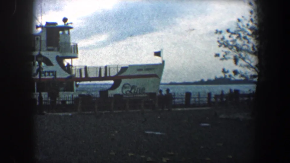 A white ferry marked with visible lettering sits at a New York City dock under a cloudy sky in soft 8mm footage.