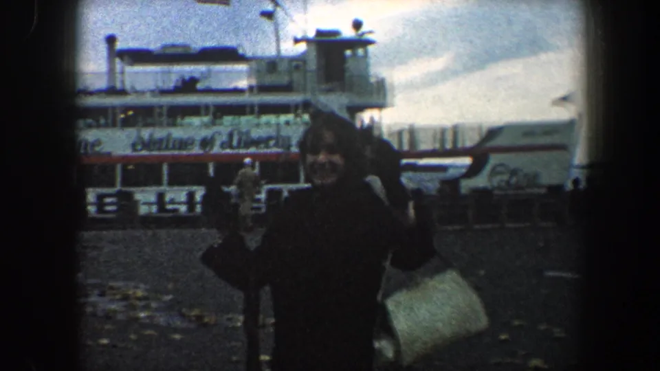 In 1960 New York City, a woman waves toward the camera beside a ferry marked Statue of Liberty Ferry.