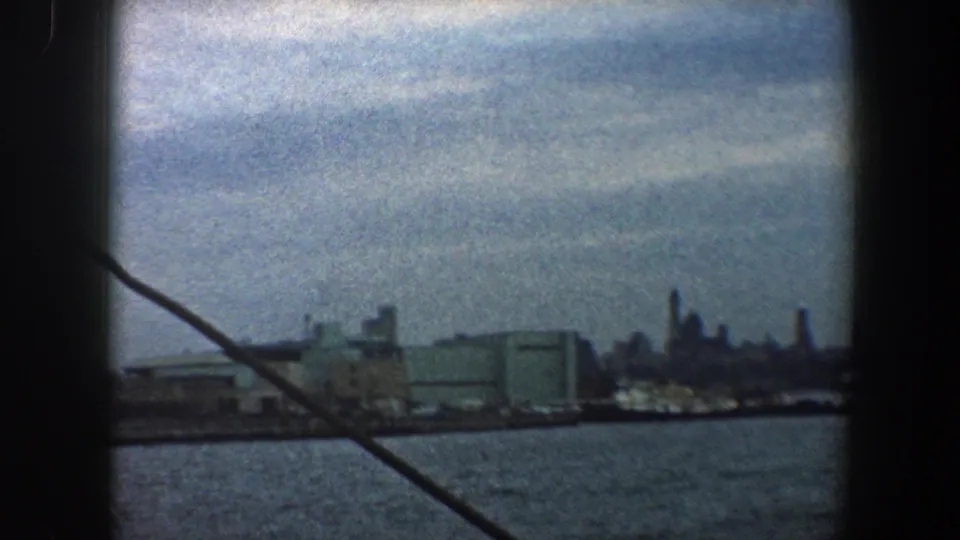 A 1960 New York harbor view shows industrial waterfront buildings and skyline silhouettes beyond the water.