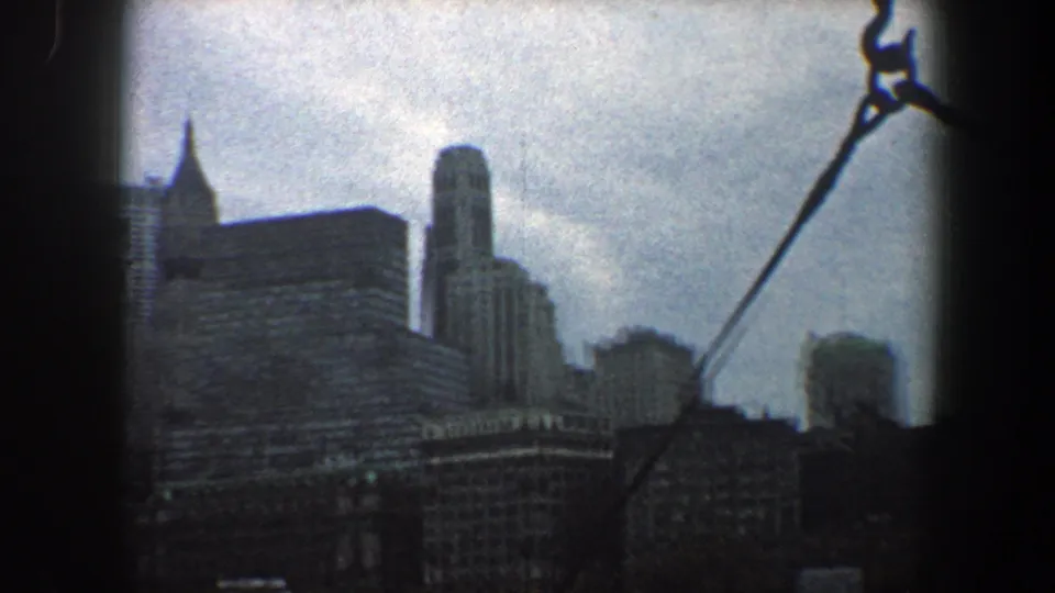 New York City skyline from a moving boat in 1960, showing towers, river water, and a diagonal cable in the foreground.