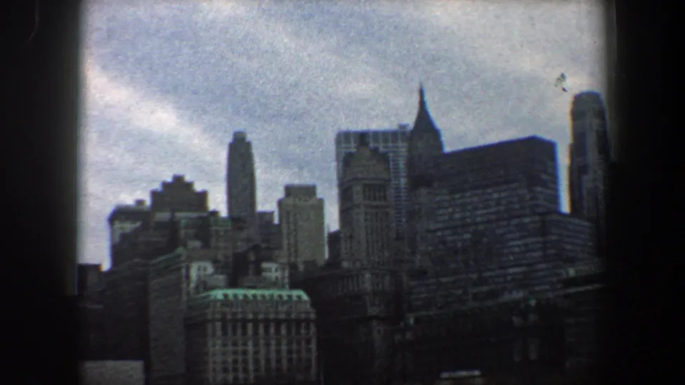 New York City skyline in 1960 8mm footage, showing dense high-rises and a prominent spired tower shape under a blue-gray sky.