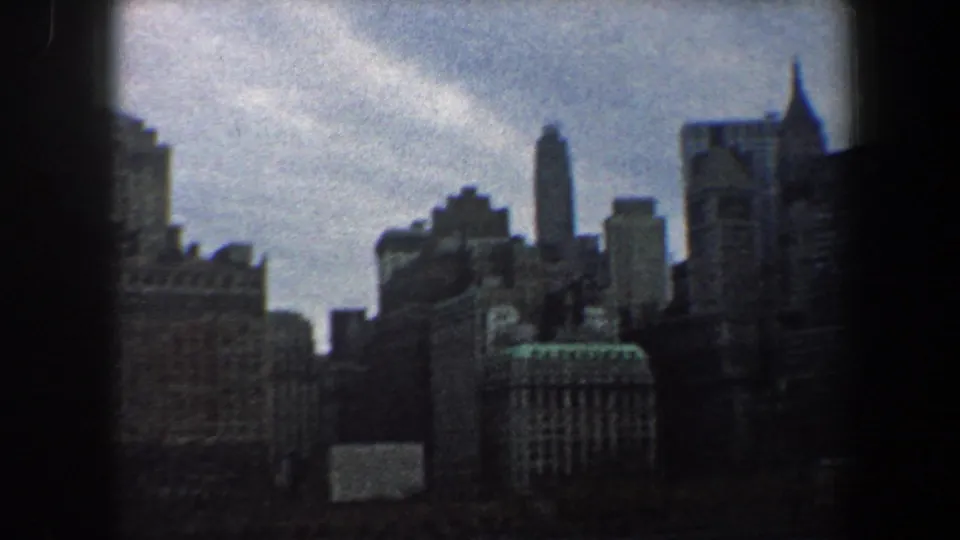Grainy 1960 home-movie frame of New York City skyscrapers, with a tall narrow tower and dark 8mm border visible.