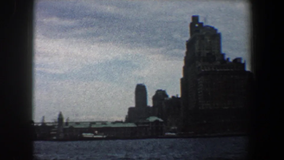 The pan reaches a wider waterfront view, with open water and dark New York City buildings against a cloudy sky.
