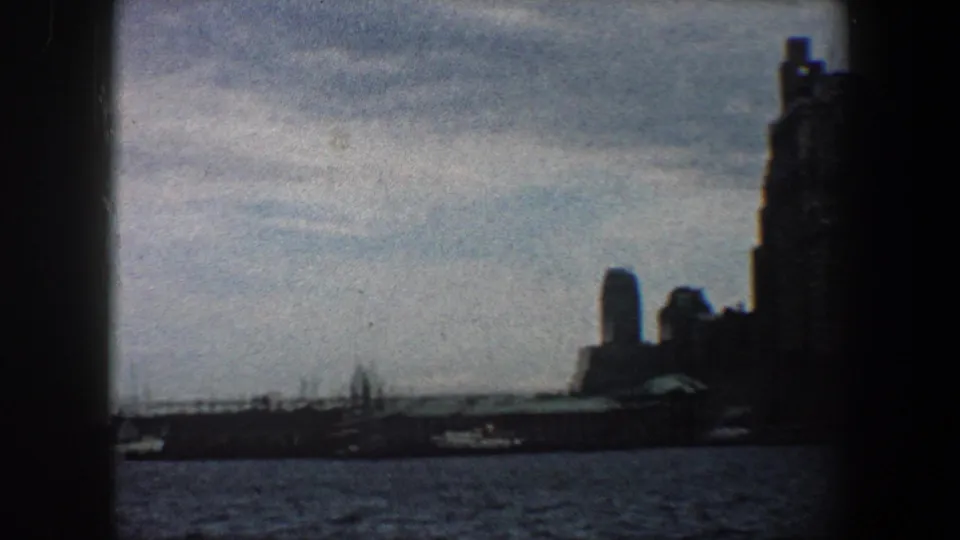 1960 8mm view of New York City buildings silhouetted across rippling harbor water, framed by dark scan borders.