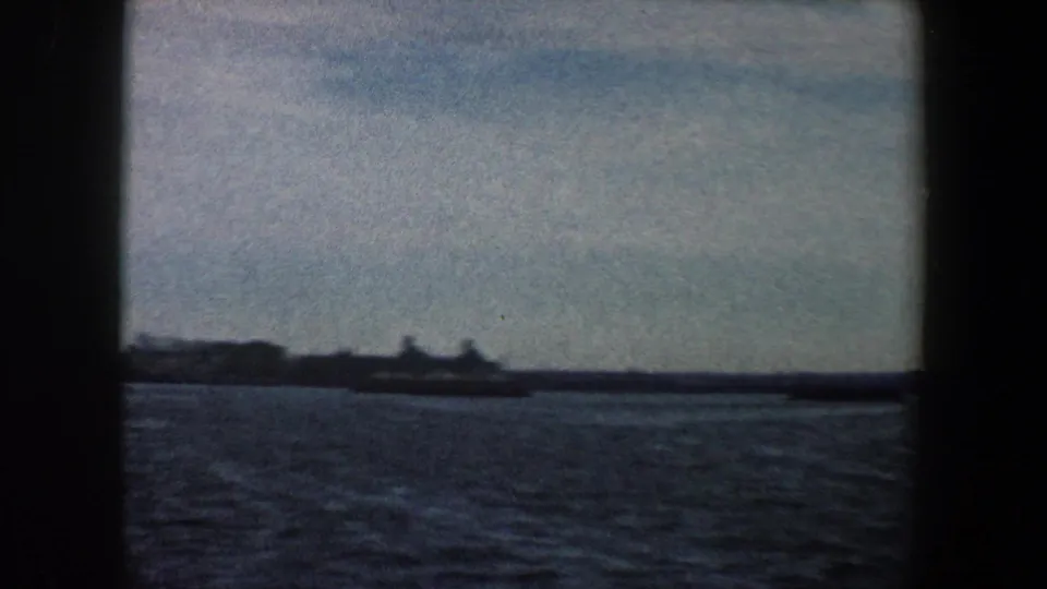 1960 New York harbor view with open water in the foreground and a distant industrial shoreline beneath a pale sky.