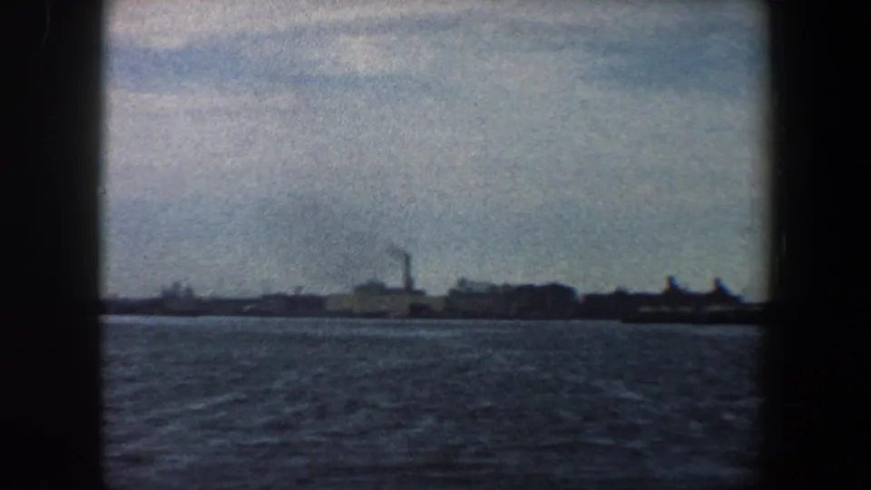 1960 8mm harbor frame showing rippling water, distant waterfront buildings, and smokestack silhouettes in New York City.
