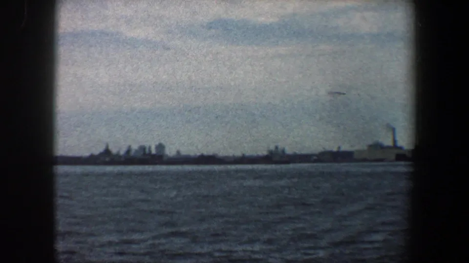 Wide 1960 New York harbor view with water in the foreground and distant buildings, smokestacks, and waterfront structures on the horizon.