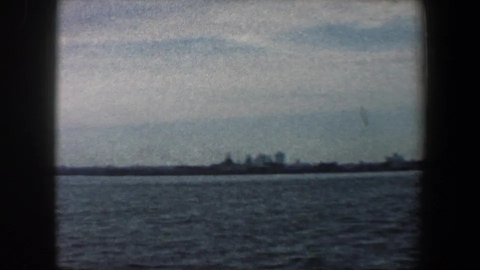 1960 8mm New York Harbor view with distant low buildings across rippled water and a hazy cloudy sky.