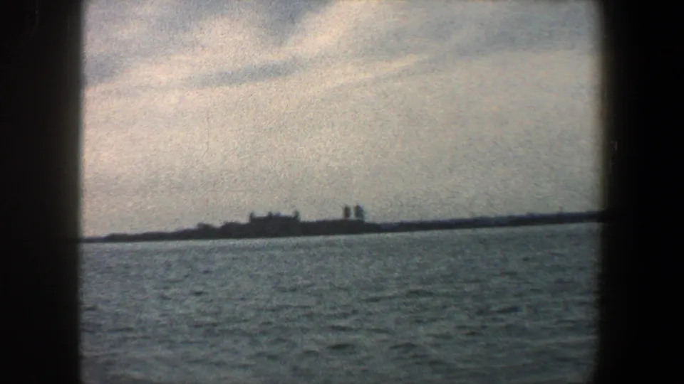 Likely Ellis Island buildings appear in silhouette across New York Harbor on grainy 1960 8mm film.