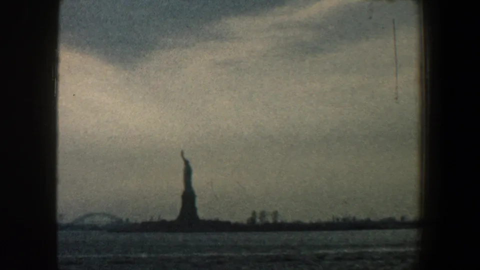 1960 8mm frame of the Statue of Liberty and low New York Harbor shoreline buildings seen from a moving vessel.