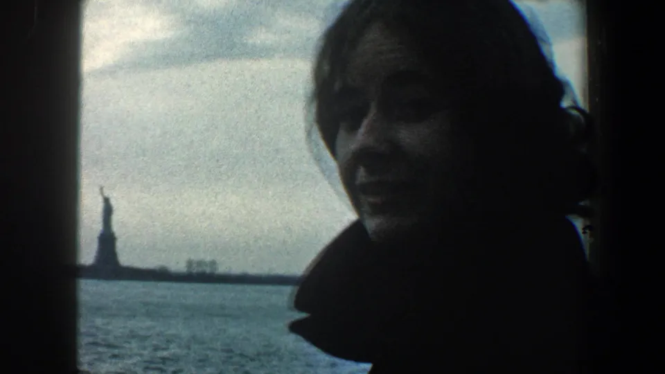 In 1960 New York City footage, a young woman in silhouette stands by a dark window frame while the Statue of Liberty appears across cloudy harbor water.