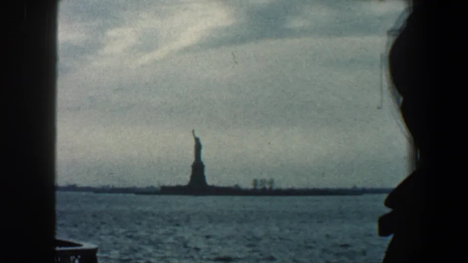 The Statue of Liberty stands across the harbor in 1960 New York City footage, framed by dark window edges and a partial silhouetted figure at right.