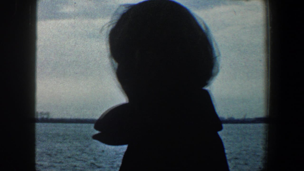 1960 New York City home-movie frame with a silhouetted woman on a boat facing the distant Statue of Liberty across the harbor.