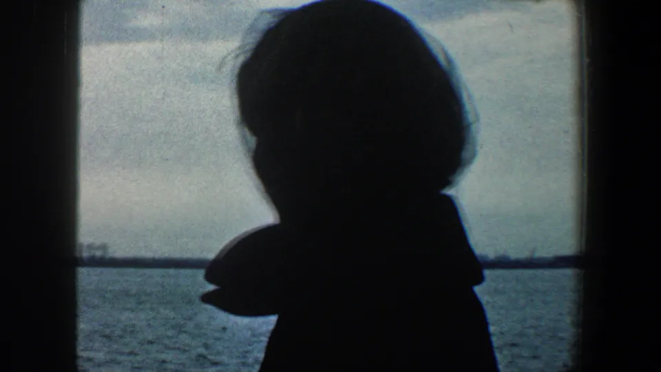 1960 New York City home-movie frame with a silhouetted woman on a boat facing the distant Statue of Liberty across the harbor.