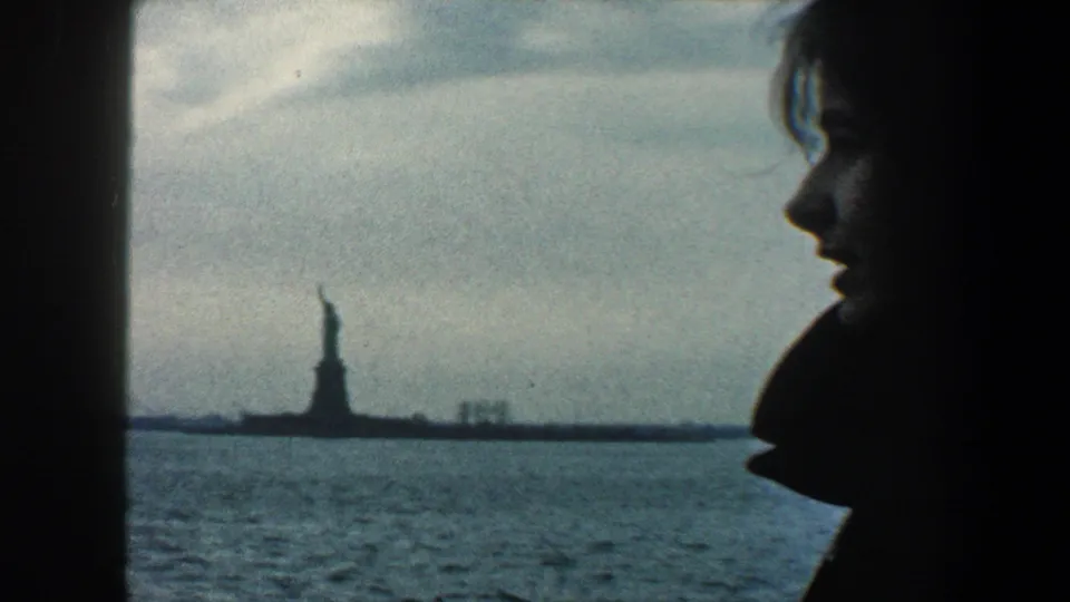 1960 New York City harbor view from a boat, with a woman in profile and the Statue of Liberty visible beyond the water.