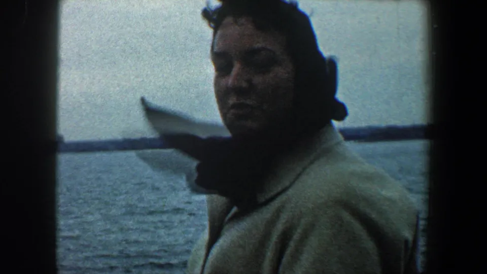 1960 New York City home-movie frame of a woman on a boat turning toward the camera while holding a folded paper by the harbor.