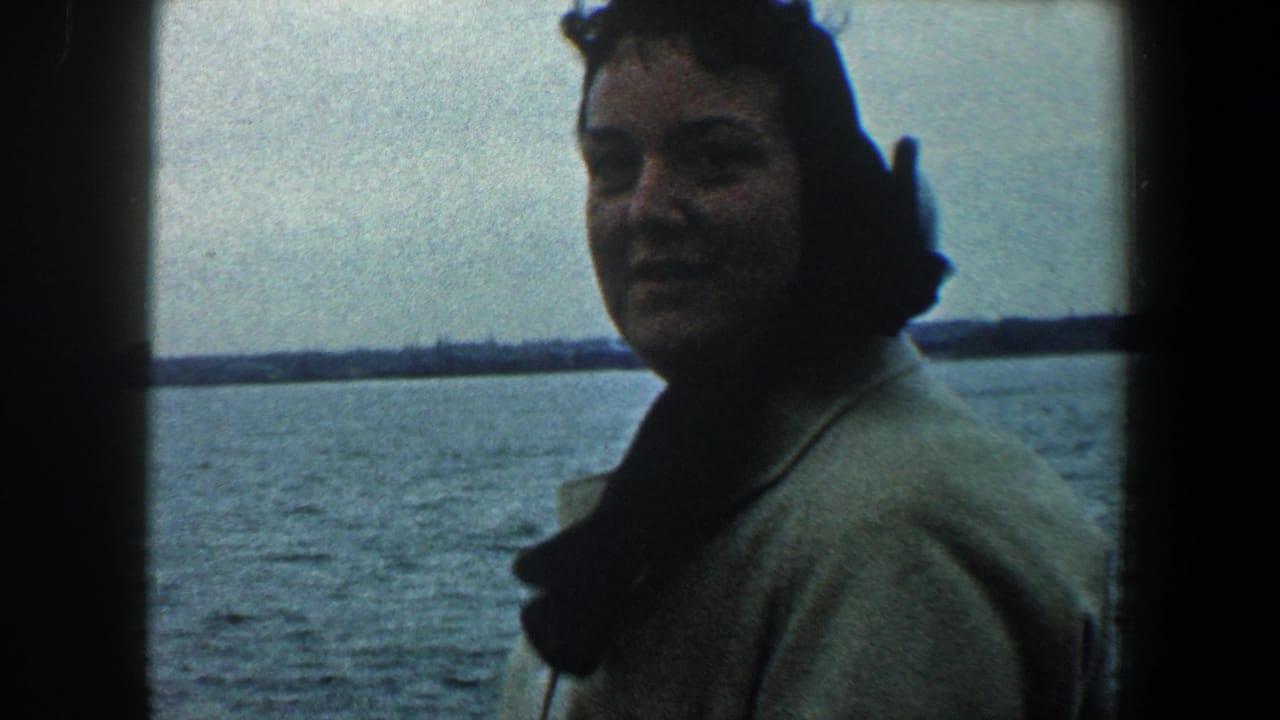 Woman in a light coat stands by a ship window with water behind her.
