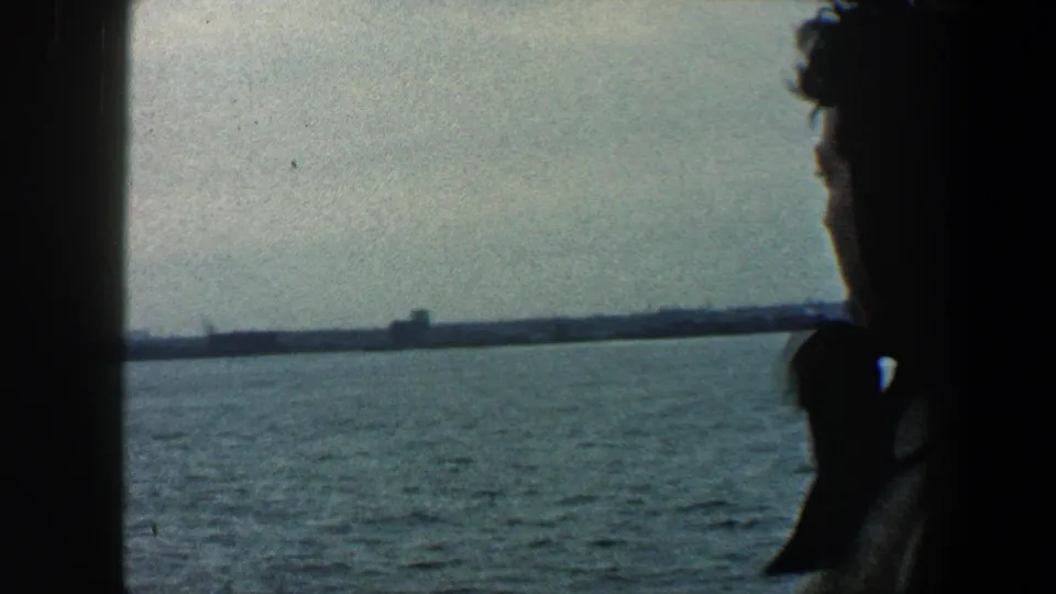 The woman is partly obscured by a dark ship window frame while open water and a distant shoreline remain visible beyond her.