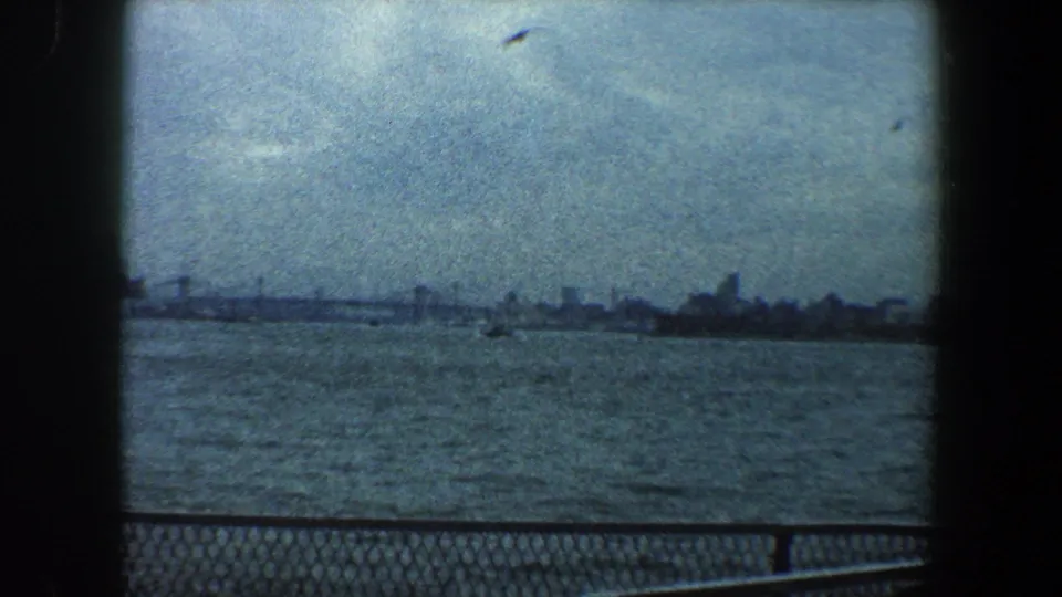 1960 8mm view of the New York City skyline across water, framed by dark film edges and a chain-link railing.