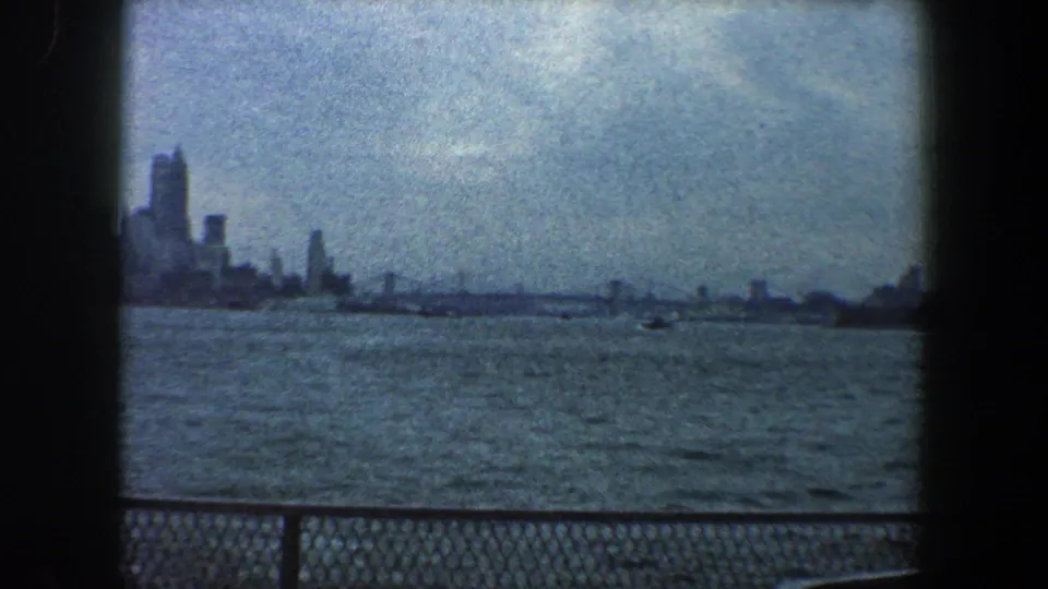 1960 New York City waterfront view showing a darker skyline section, open water, and chain-link railing.