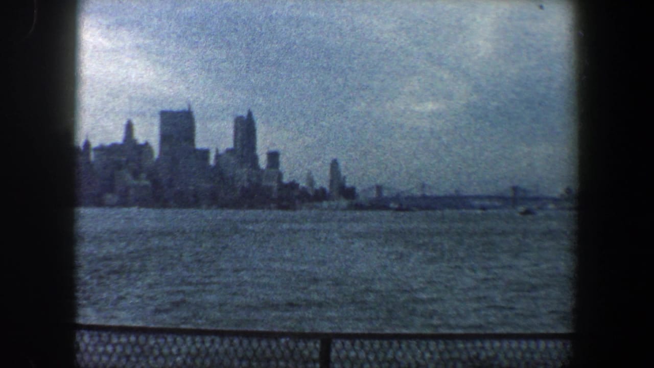 1960 8mm view of the New York City skyline seen across water, with a cloudy sky and chain-link railing in the foreground.