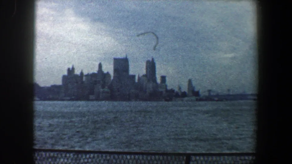 Distant 1960 New York City skyline across choppy water, framed by visible 8mm gate edges and a cloudy blue-gray sky.