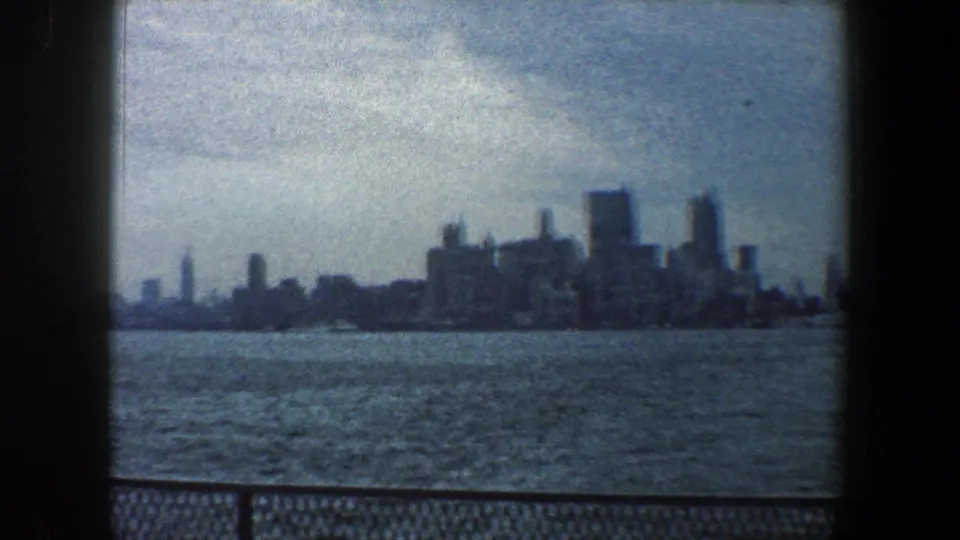 Soft 1960 New York City skyline view across water, with blue-gray cloud cover, foreground railing, and 8mm frame edges.