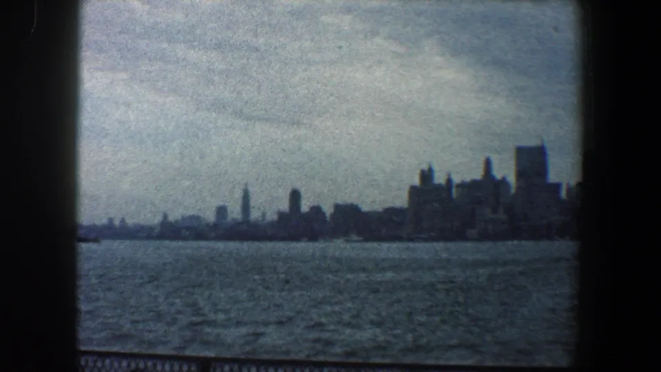 1960 8mm view of the New York City skyline seen across dark, choppy harbor water from behind a low railing.