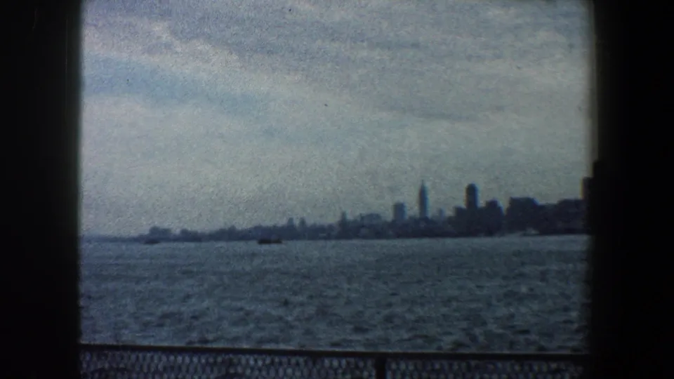 A 1960 8mm home-movie frame shows New York City buildings across choppy water, with a dark railing visible along the lower frame.