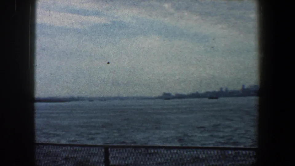 1960 8mm home-movie view of New York City across choppy harbor water, framed by dark film borders.