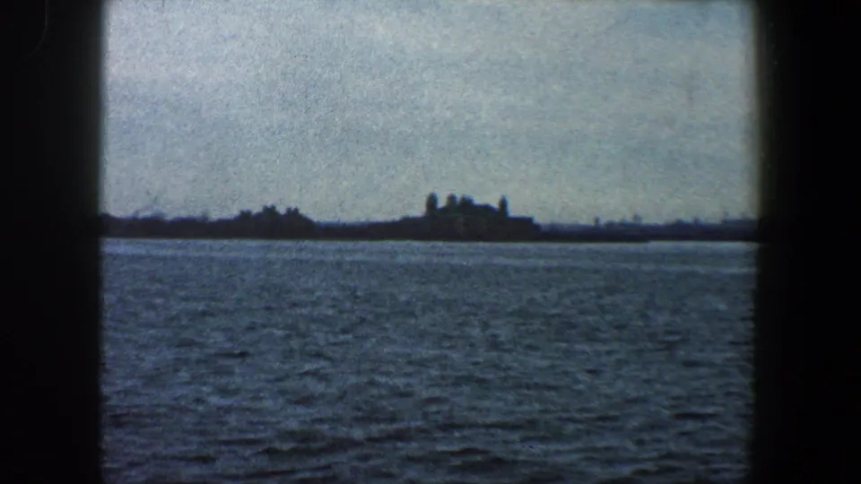 1960 home-movie frame showing a low New York City shoreline and clustered structures across rippling water.