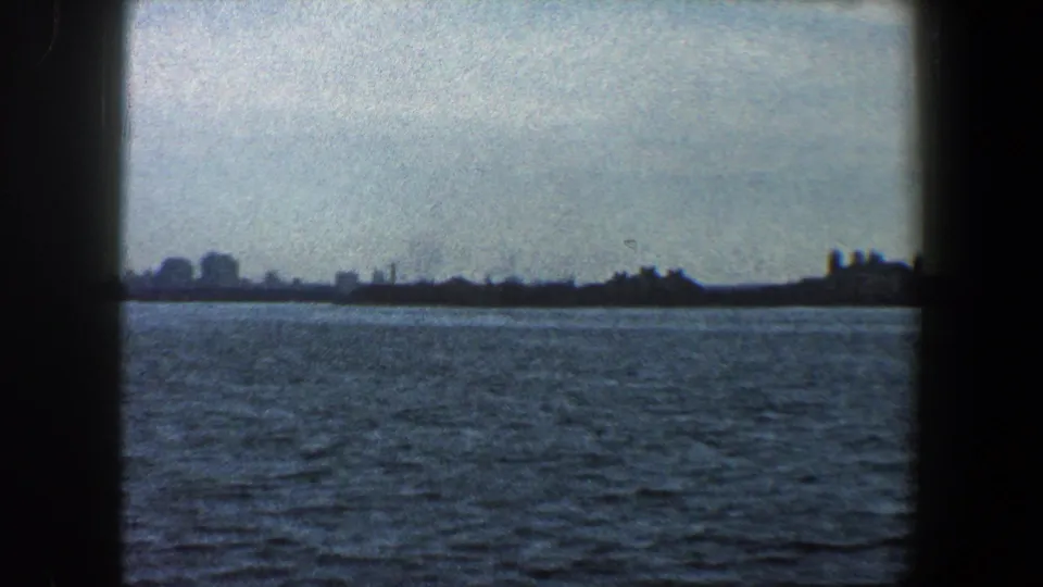 New York City waterfront in a 1960 8mm frame, seen as a distant skyline across rough harbor water.