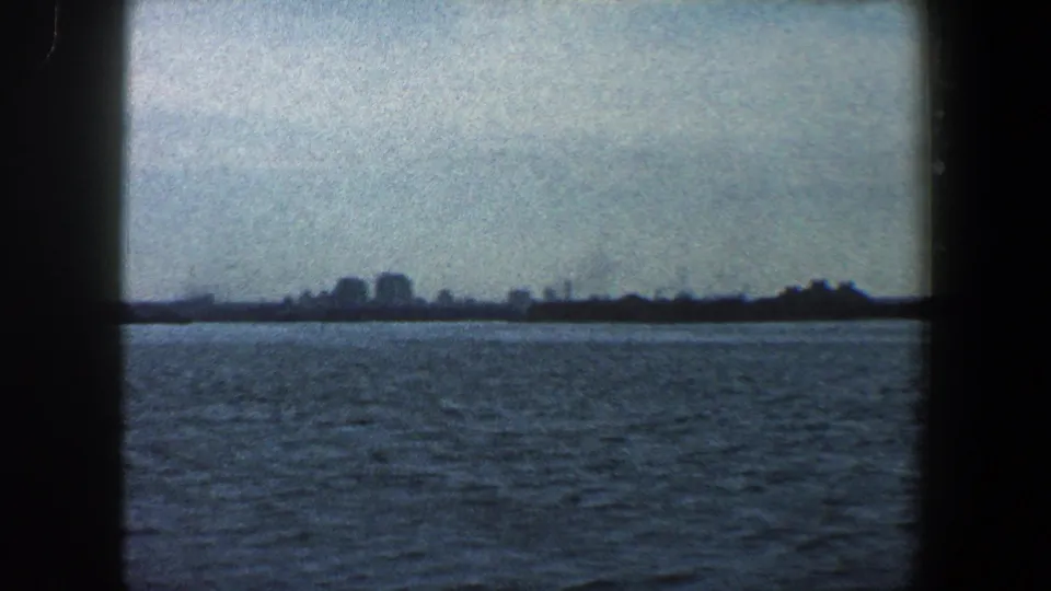 1960 New York City view across water with a hazy distant shoreline, low buildings, and dark 8mm frame borders.