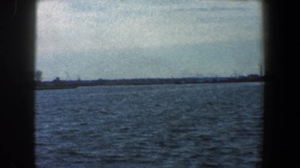 1960 New York City shoreline seen from water, with a low horizon, pale sky, and grainy 8mm film texture.
