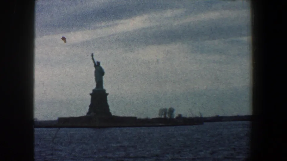 1960 8mm New York City harbor frame showing the Statue of Liberty left of center, seen from the water with shoreline and overcast sky.