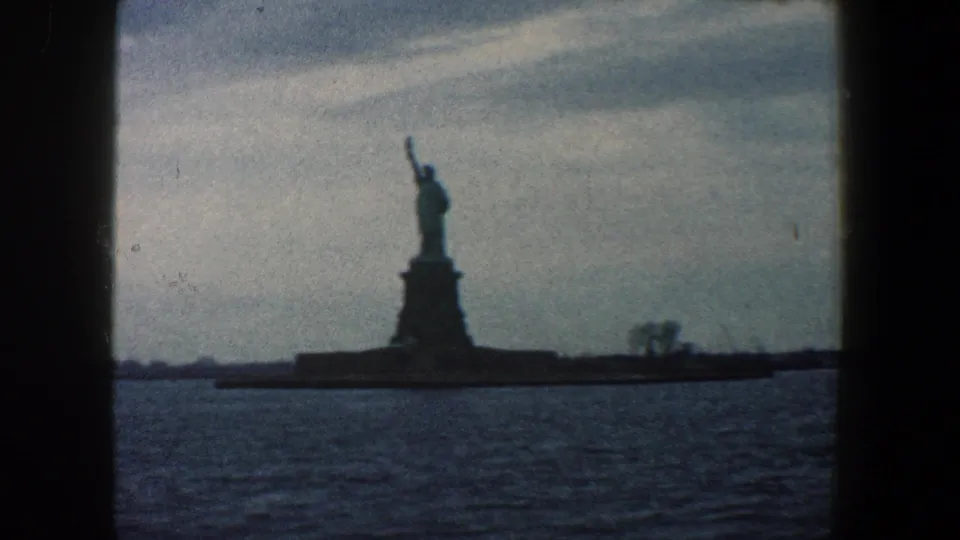 1960 8mm harbor view of the Statue of Liberty centered against a cloudy sky, with rippling water and Liberty Island shoreline visible.
