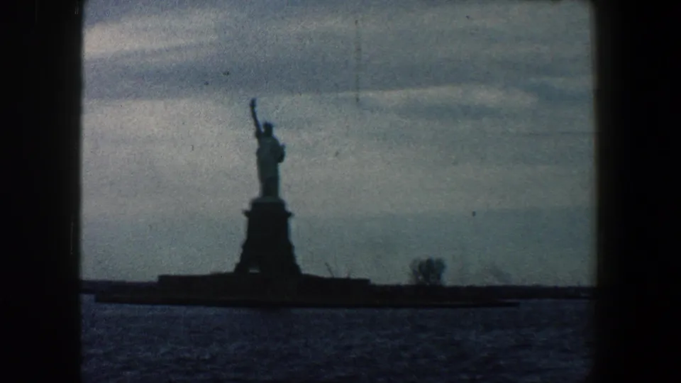 1960 8mm view of the Statue of Liberty from New York harbor, with the monument dark against a pale sky and rippled water in front.
