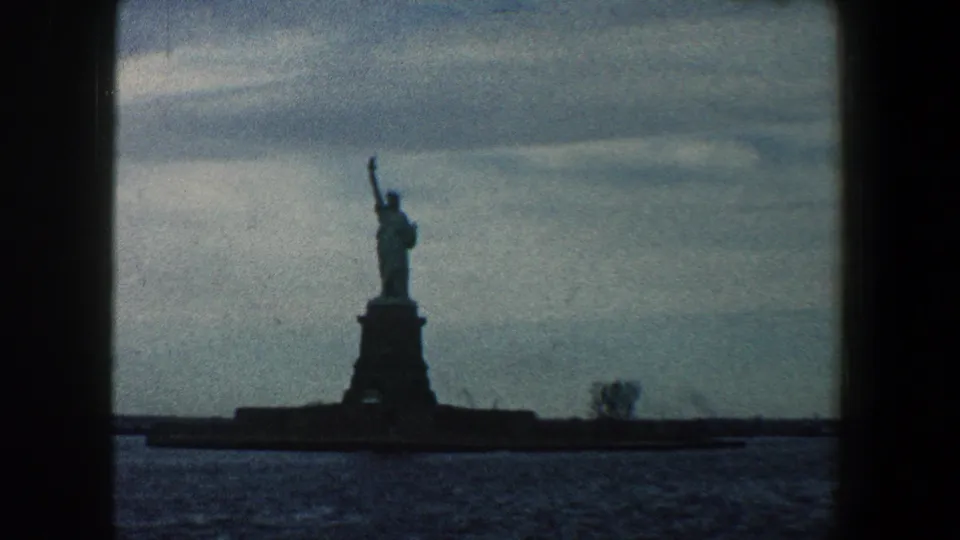 The Statue of Liberty stands centered in a 1960 New York harbor home-movie frame, with overcast sky, dark water, and visible film texture.