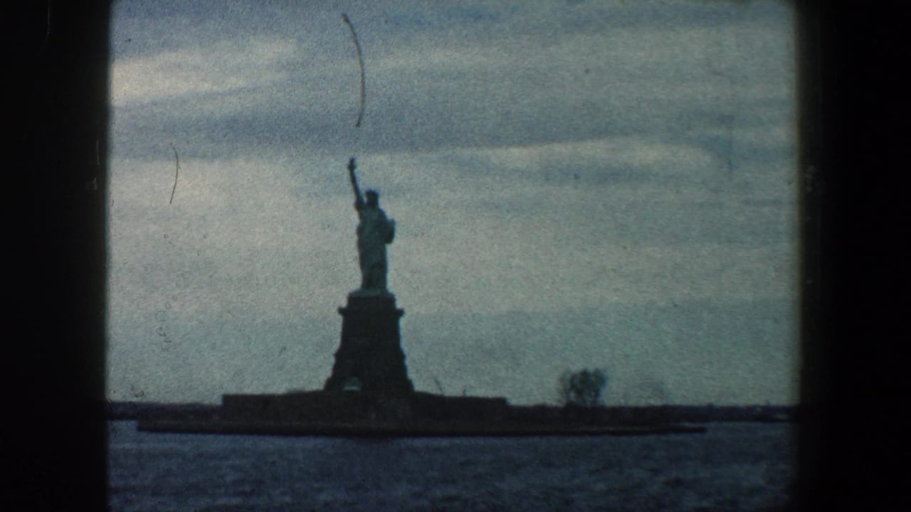 Distant Statue of Liberty silhouetted across harbor water under a cloudy sky.