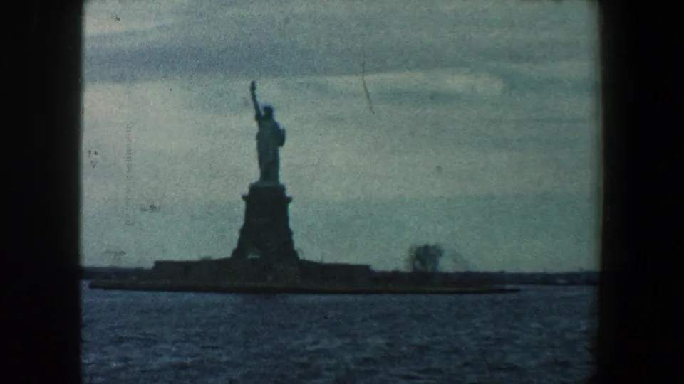 1960 New York travel footage frame showing the Statue of Liberty across the harbor, with the monument silhouetted and film grain visible.