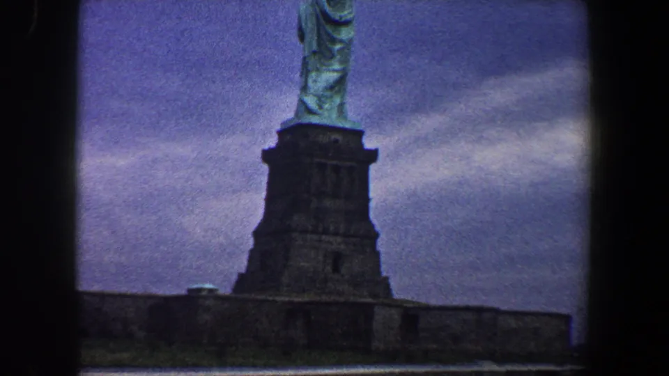 A lower angle from the 1960 home movie shows the Statue of Liberty, its pedestal, and a base wall beneath a blue-purple overcast sky.