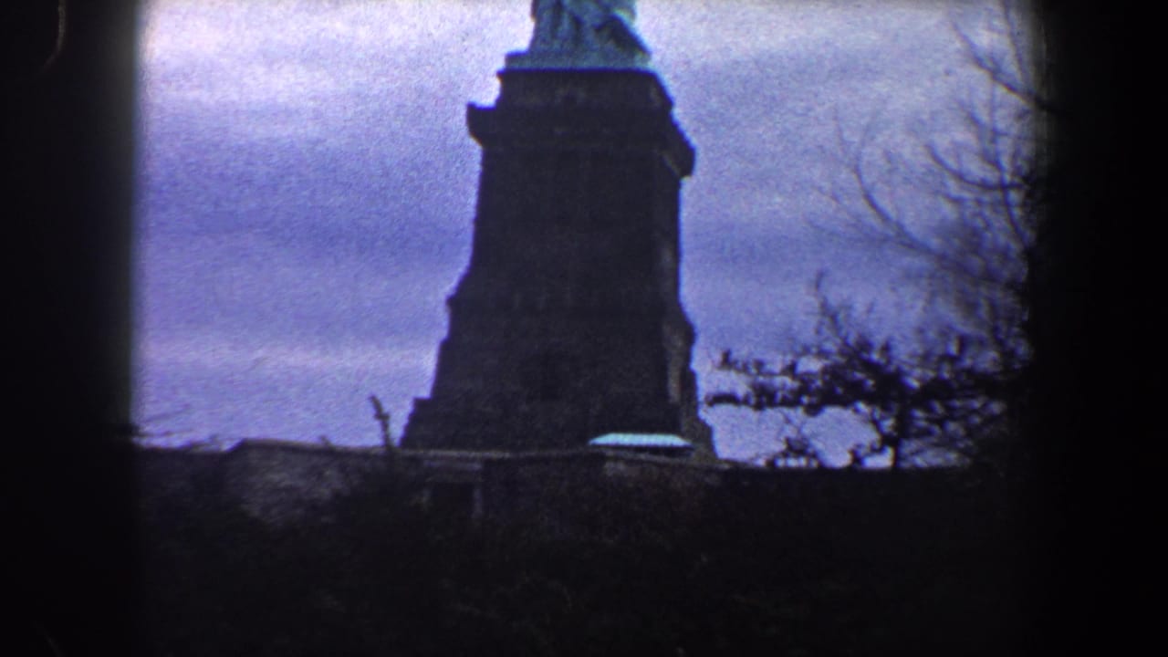 1960 New York City home-movie frame showing the Statue of Liberty pedestal from a low angle with bare branches and cloudy sky.