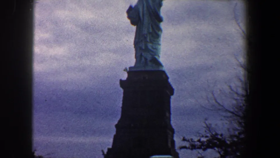 Low-angle 8mm view of the Statue of Liberty rising above its pedestal, with bare branches against a cloudy New York sky.