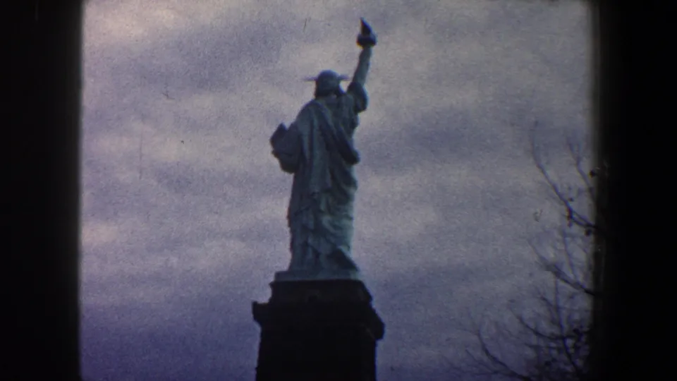 1960 New York City home-movie frame showing a fuller low-angle view of the Statue of Liberty and torch under a clouded sky.