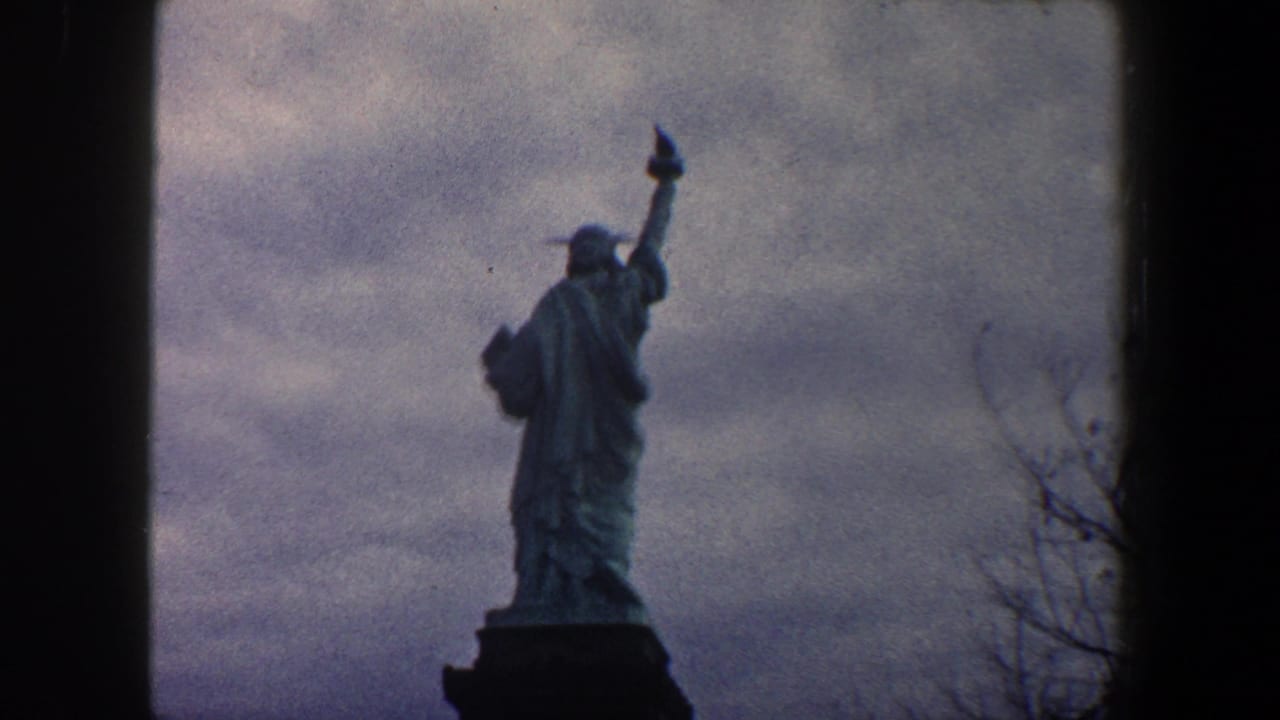 1960 New York City 8mm frame showing the Statue of Liberty from a rear low angle, silhouetted against an overcast sky.
