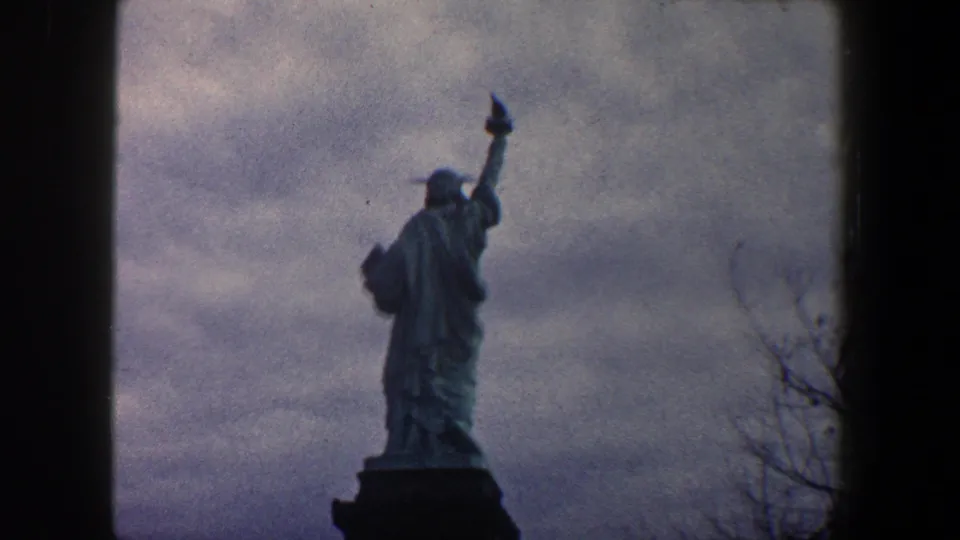 1960 New York City 8mm frame showing the Statue of Liberty from a rear low angle, silhouetted against an overcast sky.