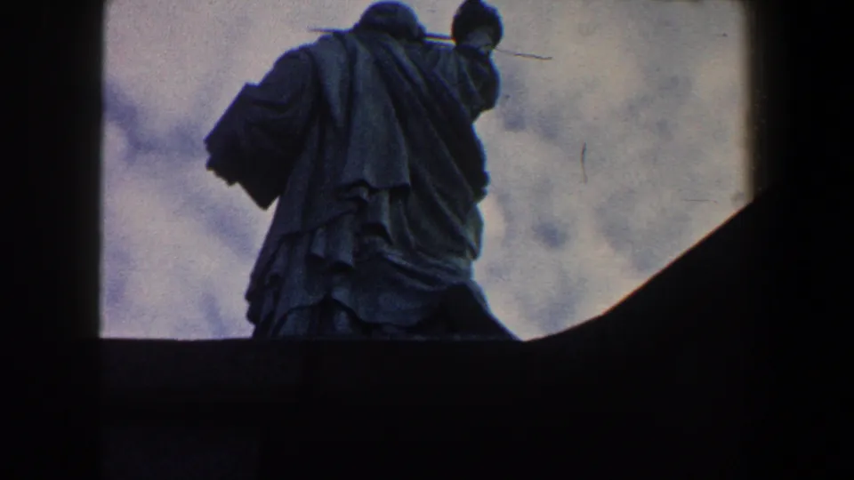 Close 8mm frame of the Statue of Liberty from behind in New York City, with robes and raised arm against gray clouds.
