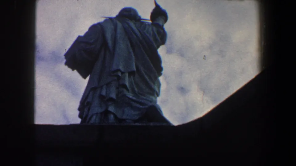1960 8mm low-angle view of the Statue of Liberty's back, with draped robes and raised torch against a muted cloudy sky.